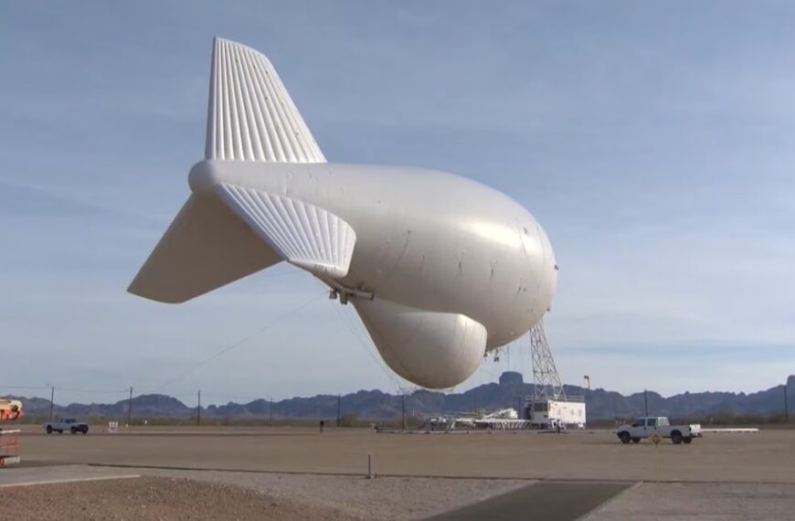 Large white airship inflated on a runway, tethered to a support truck and scaffolding with desert mountains in the background.