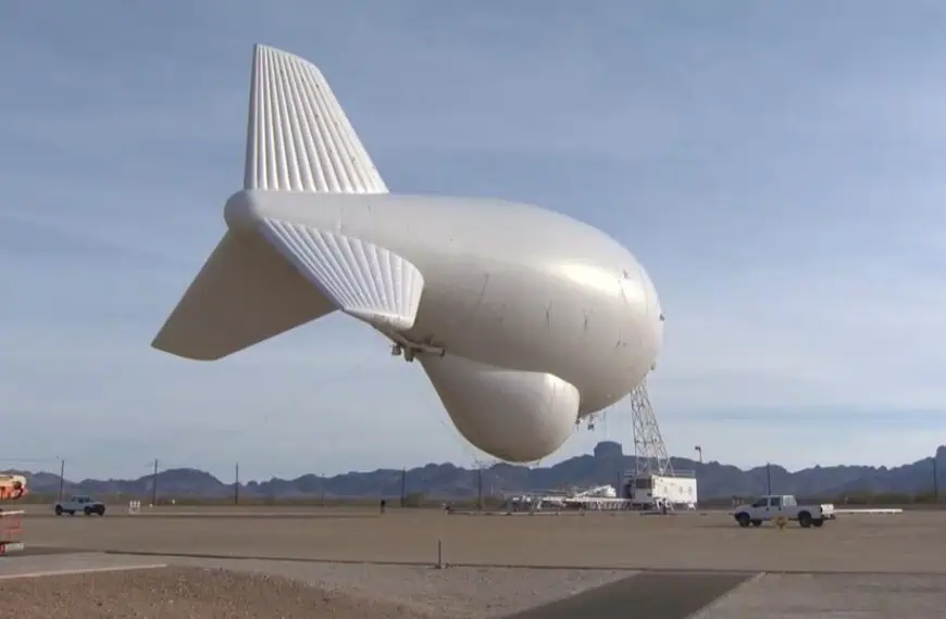 Large white airship inflated on a runway, tethered to a support truck and scaffolding with desert mountains in the background.