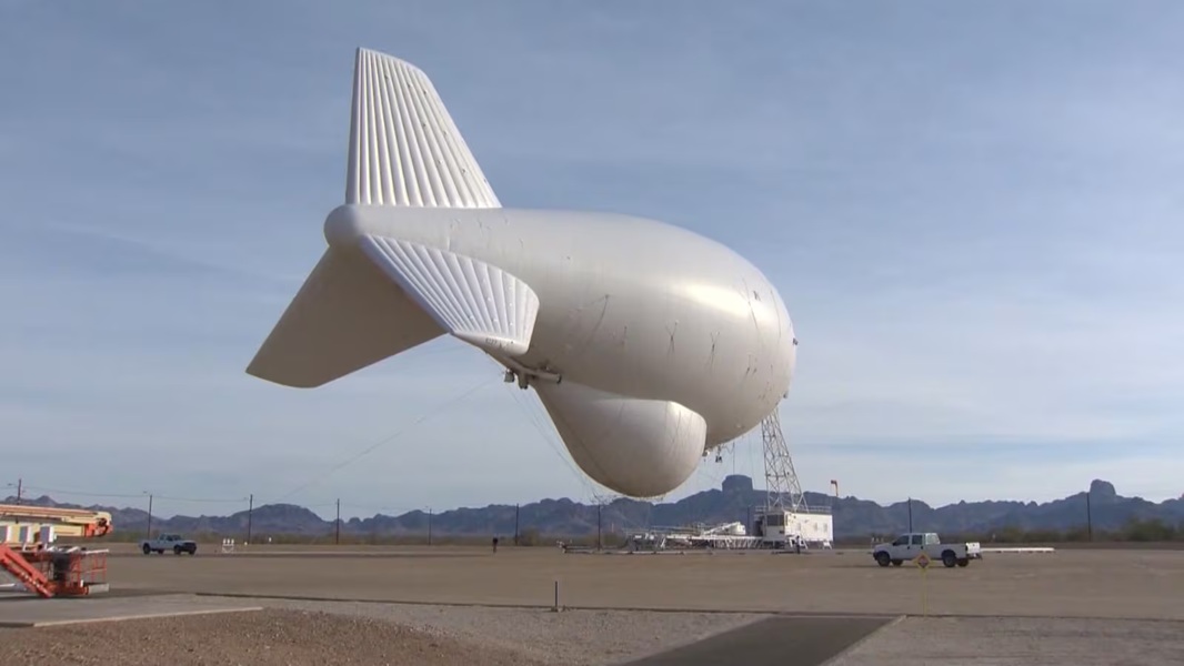 Large white airship inflated on a runway, tethered to a support truck and scaffolding with desert mountains in the background.