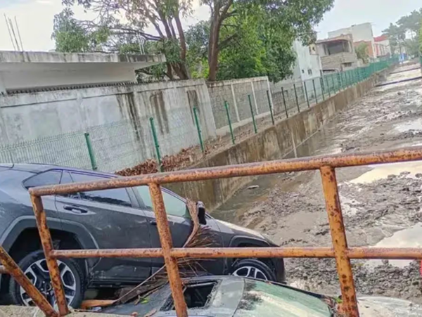 Gray SUV wedged against a rusted railing over a deep muddy ditch in a residential alley; a damaged car blocks part of the pathway.