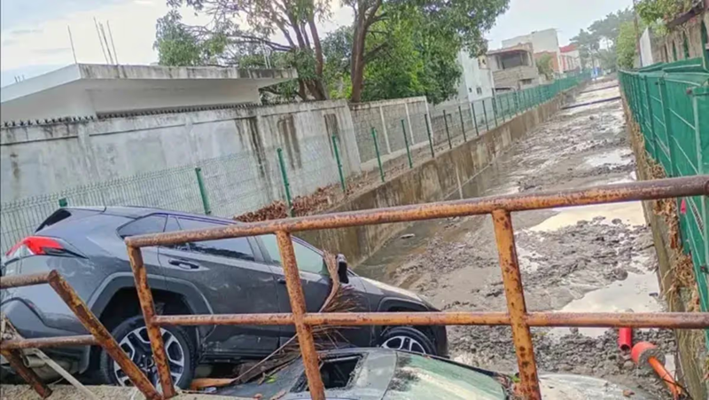 Gray SUV wedged against a rusted railing over a deep muddy ditch in a residential alley; a damaged car blocks part of the pathway.