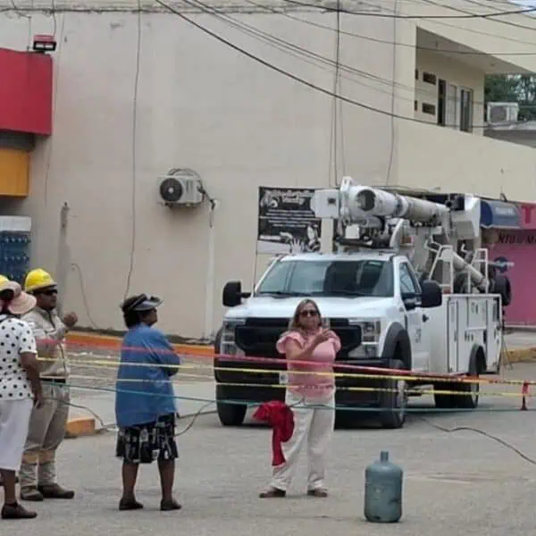 Construction workers in hard hats stand behind caution tape near a white utility bucket truck on a street with shops in the background.