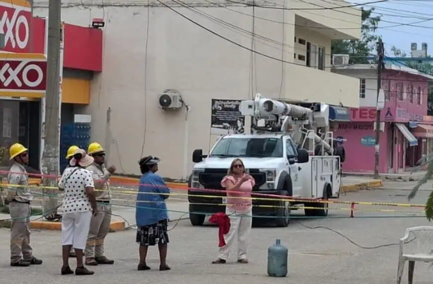 Construction workers in hard hats stand behind caution tape near a white utility bucket truck on a street with shops in the background.