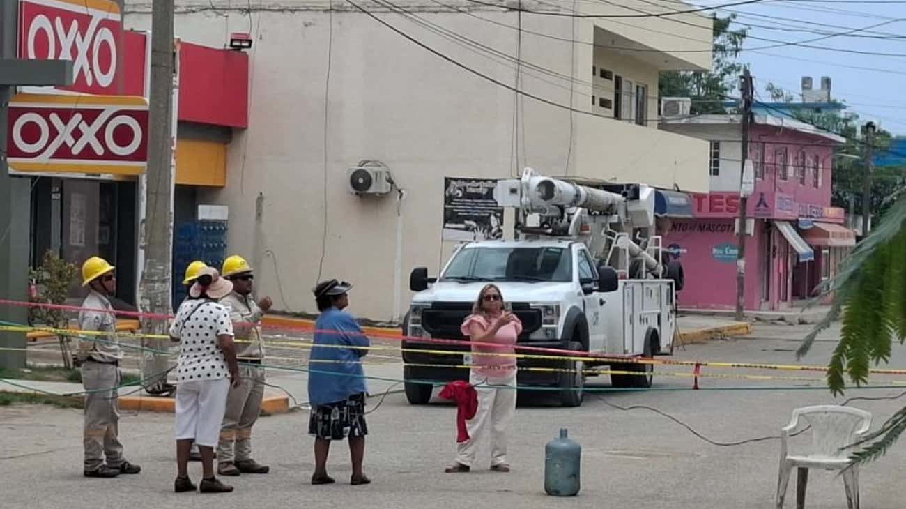 Construction workers in hard hats stand behind caution tape near a white utility bucket truck on a street with shops in the background.
