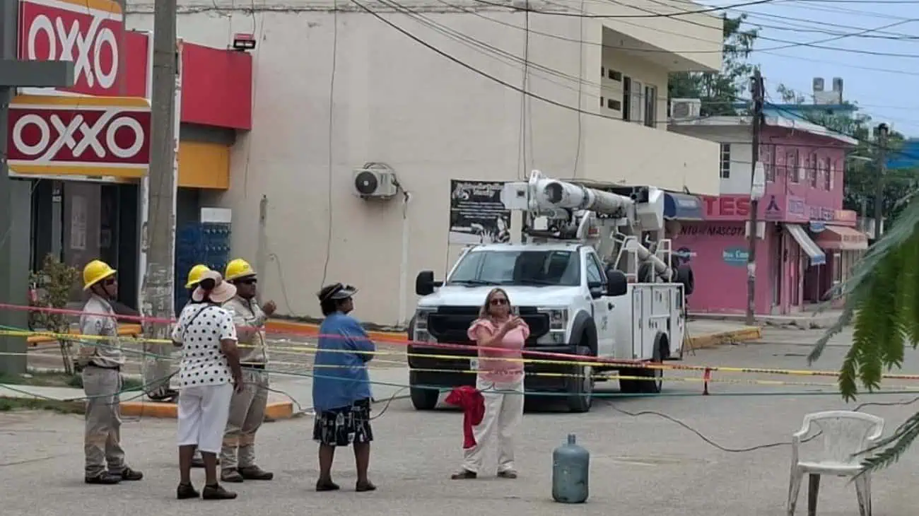Construction workers in hard hats stand behind caution tape near a white utility bucket truck on a street with shops in the background.