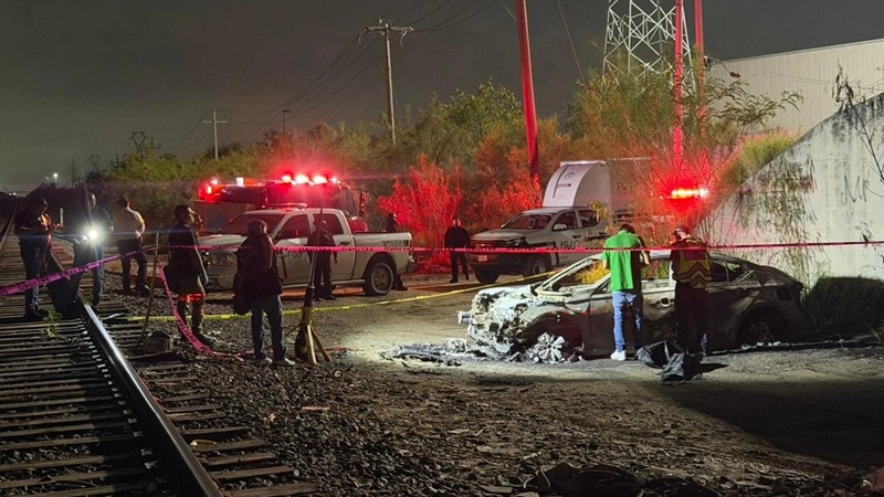 Night scene of a burned-out car by railroad tracks with emergency responders and red siren lights.