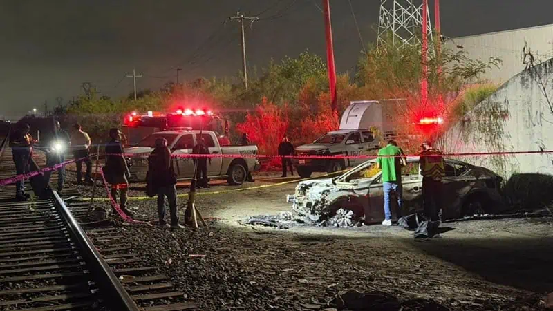 Night scene of a burned-out car by railroad tracks with emergency responders and red siren lights.