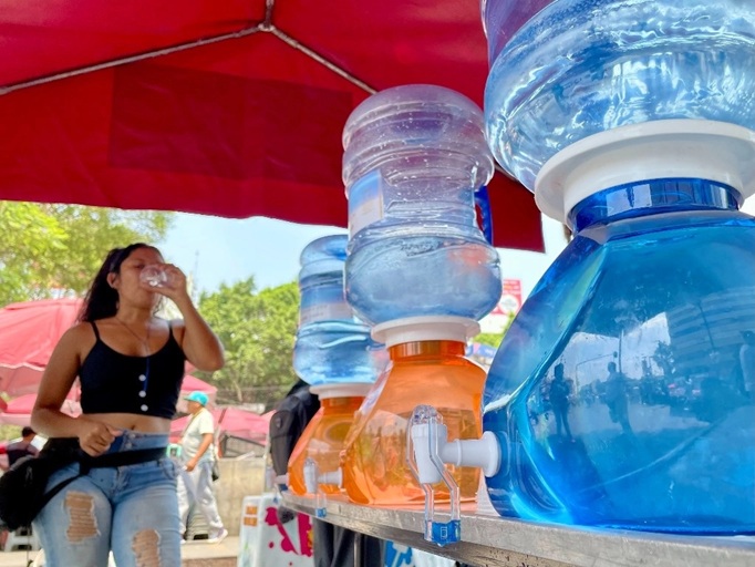 Street-side water dispenser with large blue water jugs and taps; a woman drinks from a cup in the background under a red canopy.