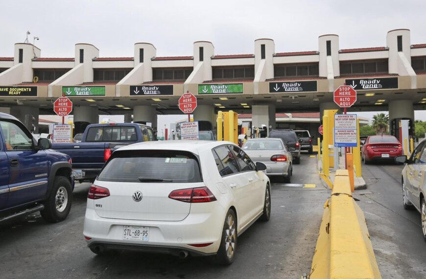 Cars line up at a toll plaza with yellow barriers and 'Ready lanes' signs above the booths; a white VW GTI is closest to the camera on the left side of the lane.