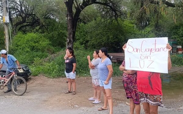 Group of women standing on a dirt road by a river, one holding a sign that reads 'Sin Luz' while a cyclist passes on the left.