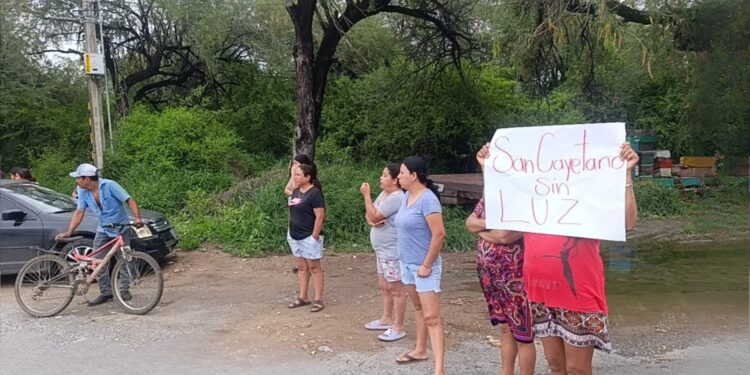 Group of women standing on a dirt road by a river, one holding a sign that reads 'Sin Luz' while a cyclist passes on the left.