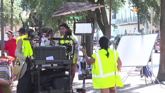 Film crew on a sunny street, wearing yellow vests, operating a large camera rig with lights and monitors.