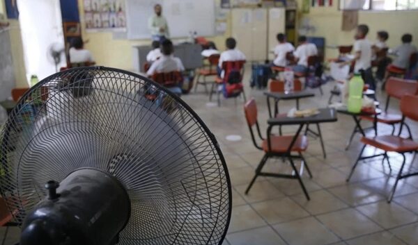 Classroom scene with students at desks and a teacher at the front; a large electric fan dominates the foreground, cooling the room.