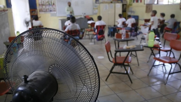 Classroom scene with students at desks and a teacher at the front; a large electric fan dominates the foreground, cooling the room.