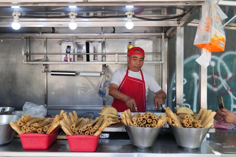 Street food vendor in a red apron cooks fried snacks behind a metal stall, with bowls of fried tubes and pastries in front.