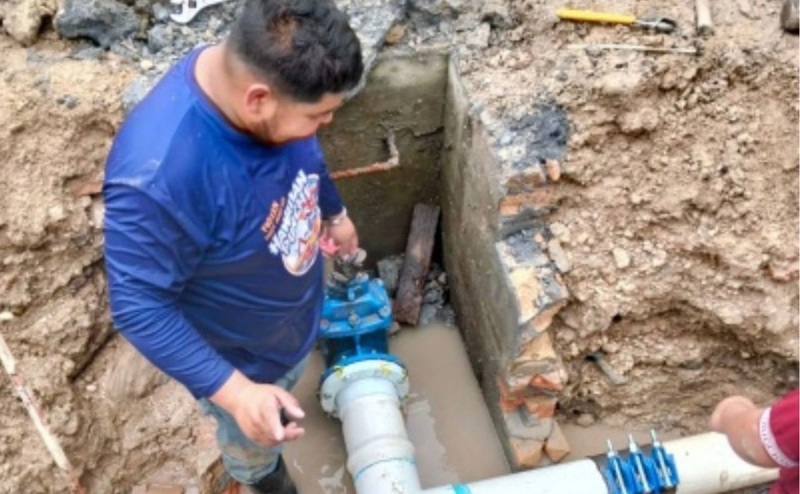 Construction worker in a blue shirt installing a PVC drainage pipe in a muddy trench.