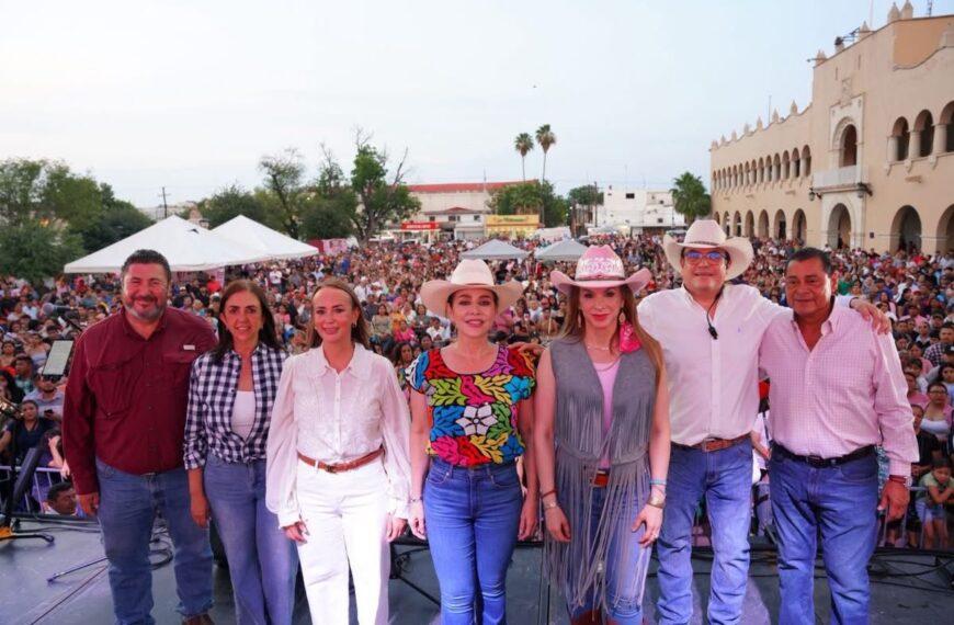 Eight people stand on a stage posing for a photo as a large festival crowd gathers behind them near a beige arched building.