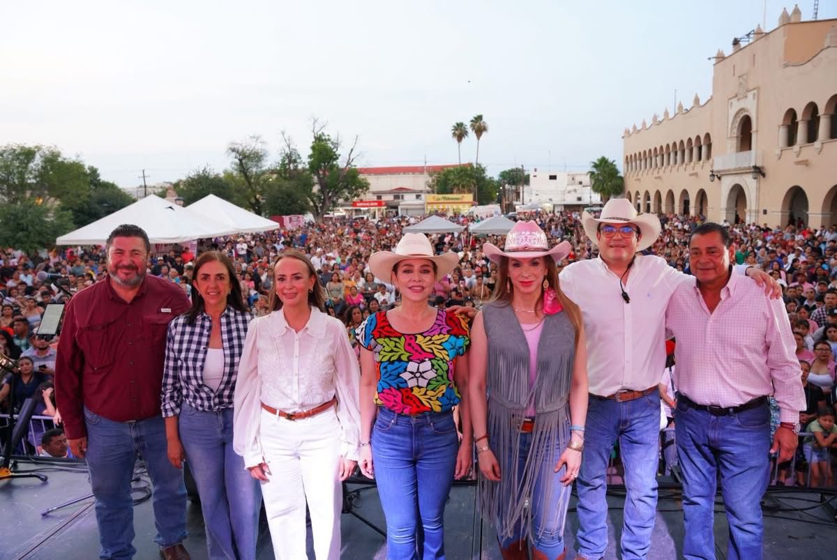 Eight people stand on a stage posing for a photo as a large festival crowd gathers behind them near a beige arched building.