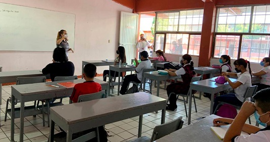 Teacher at whiteboard presenting to a classroom of students seated at desks, some wearing masks, in a bright room.