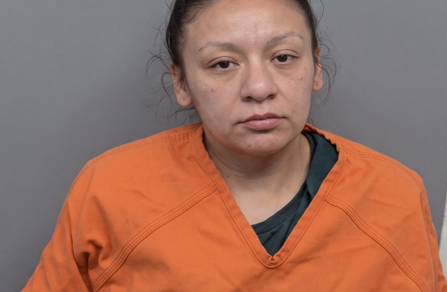 Front-facing mugshot of a woman with brown hair in a bun, wearing an orange jail jumpsuit against a gray backdrop.