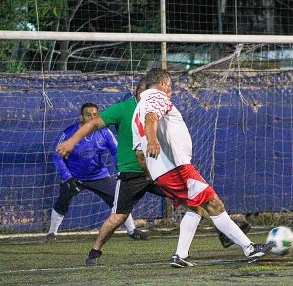 Soccer players contest the ball near the goal as a goalkeeper watches from the net on an artificial turf field. Green-shirted defender challenges a white-and-red attacker as the ball rolls toward the net.