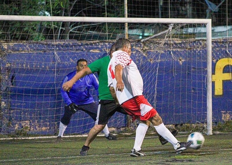 Soccer players contest the ball near the goal as a goalkeeper watches from the net on an artificial turf field. Green-shirted defender challenges a white-and-red attacker as the ball rolls toward the net.