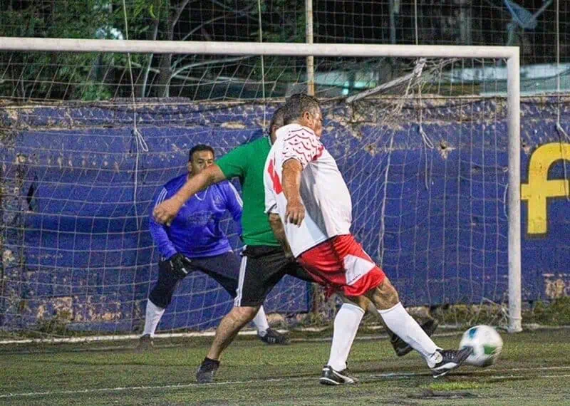 Soccer players contest the ball near the goal as a goalkeeper watches from the net on an artificial turf field. Green-shirted defender challenges a white-and-red attacker as the ball rolls toward the net.