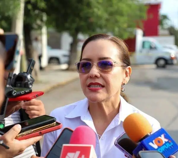 Woman wearing sunglasses speaks to reporters amid microphones outside, answering questions about a news event.