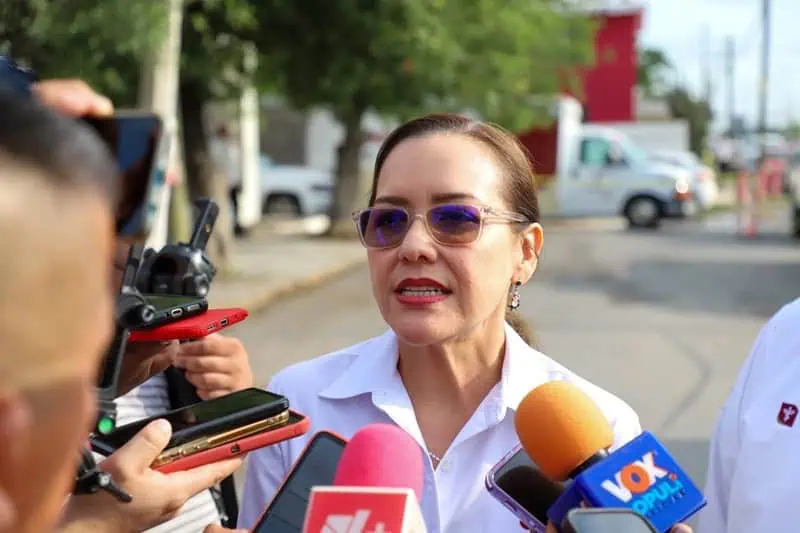Woman wearing sunglasses speaks to reporters amid microphones outside, answering questions about a news event.