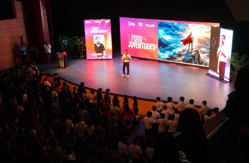 Speaker standing center stage delivering a talk at Foro Juventudes, with colorful backdrops and an audience watching from rows of seats.