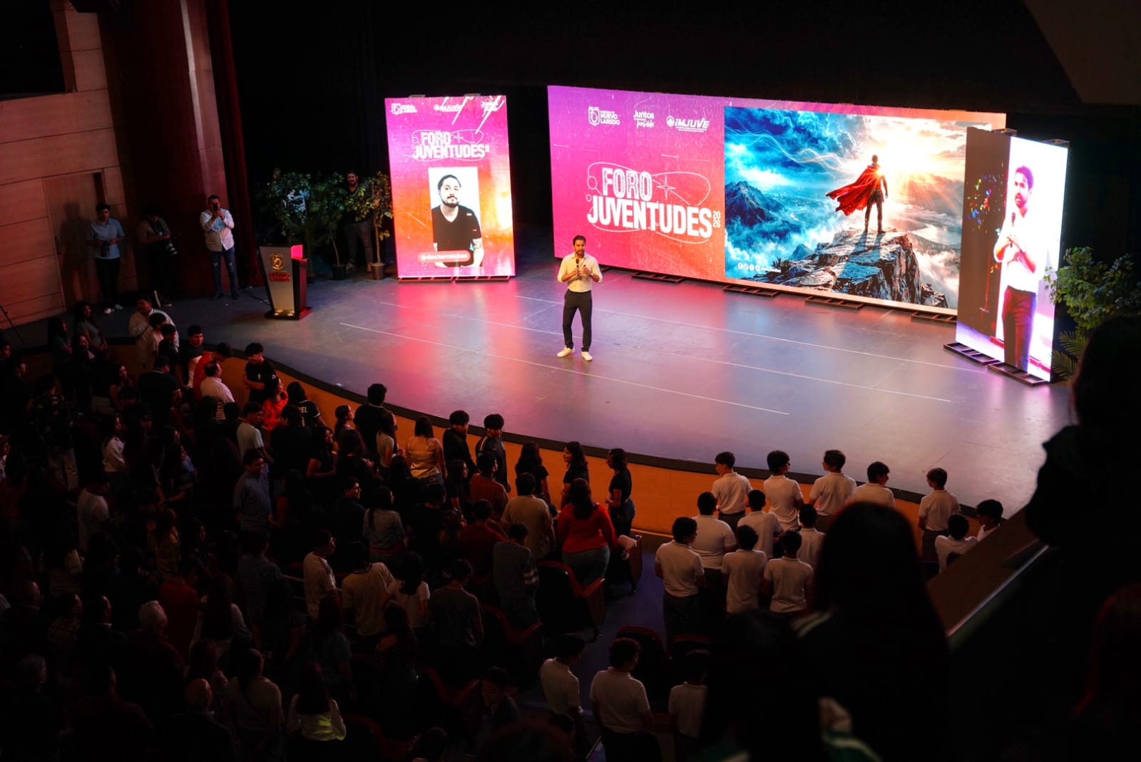 Speaker standing center stage delivering a talk at Foro Juventudes, with colorful backdrops and an audience watching from rows of seats.