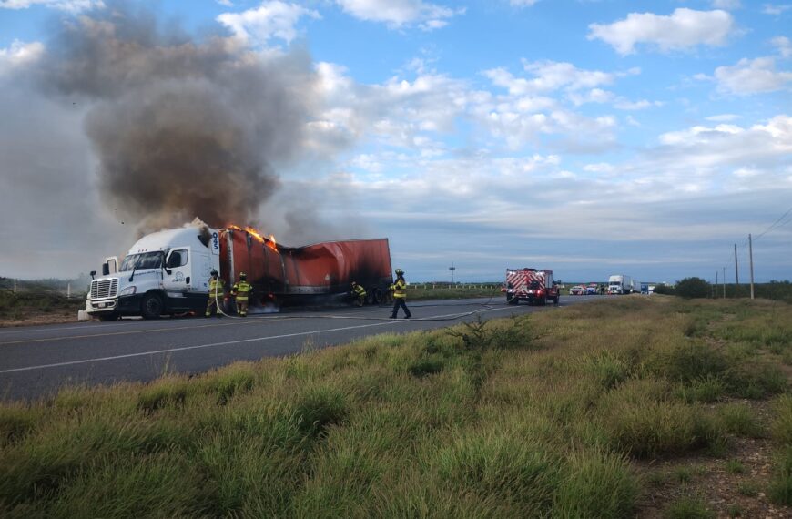 Truck and attached red trailer on fire on a highway, firefighters actively battling the blaze as flames and smoke rise behind the cab.
