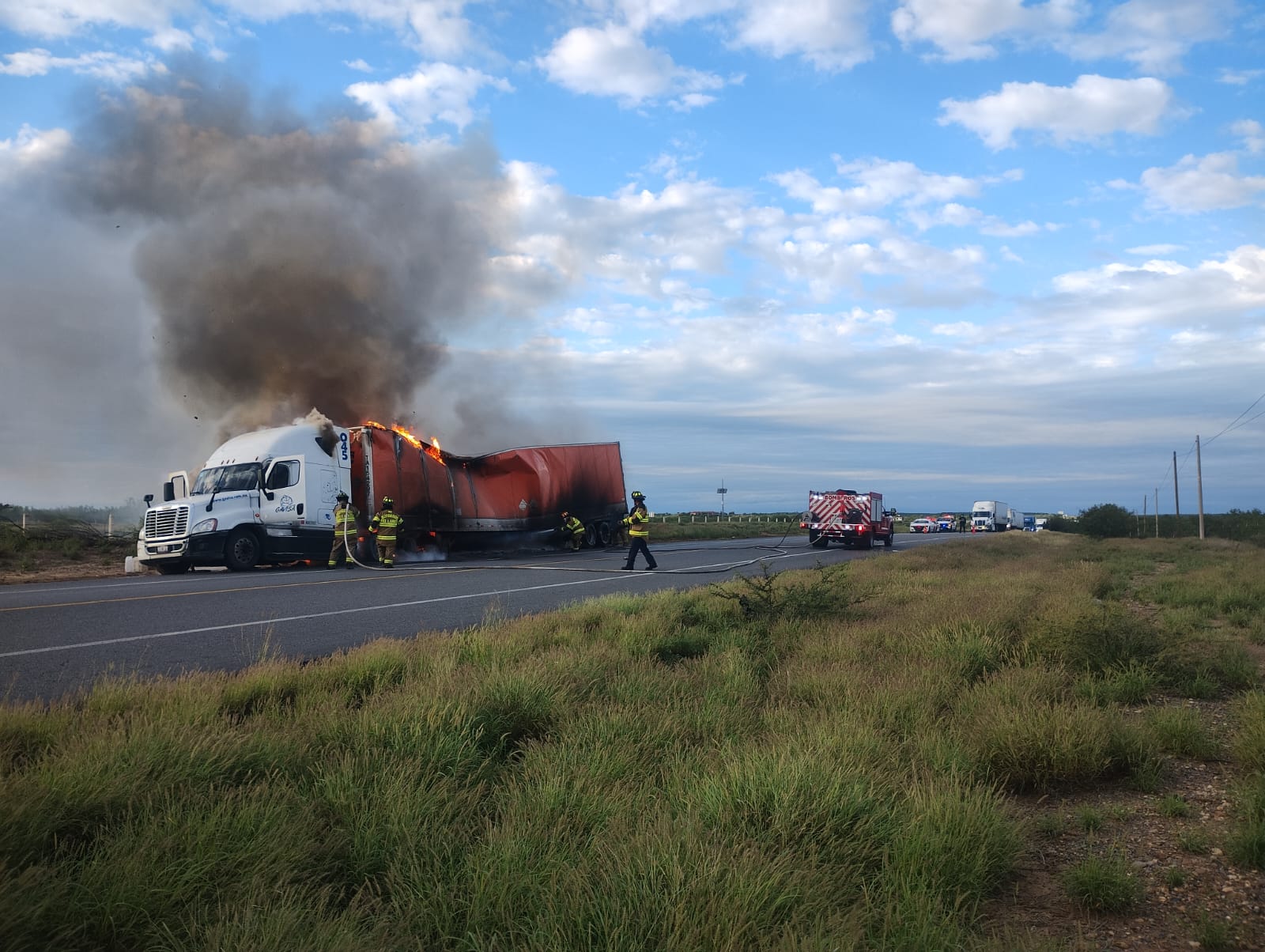 Truck and attached red trailer on fire on a highway, firefighters actively battling the blaze as flames and smoke rise behind the cab.