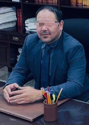 Man in a blue suit sits at a desk in an office, eyes pixelated for privacy; pencil holder and stacks of papers on the desk behind him.