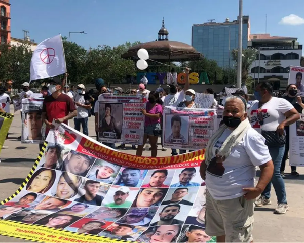 Protest in a city square with banners and posters of missing people; participants wear masks and white shirts, holding a large collage banner of faces.