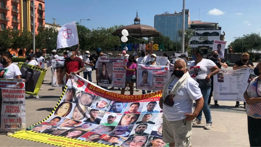 Protest in a city square with banners and posters of missing people; participants wear masks and white shirts, holding a large collage banner of faces.