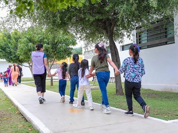 Children holding hands walk in a line along a sunny sidewalk beside trees and a white building.