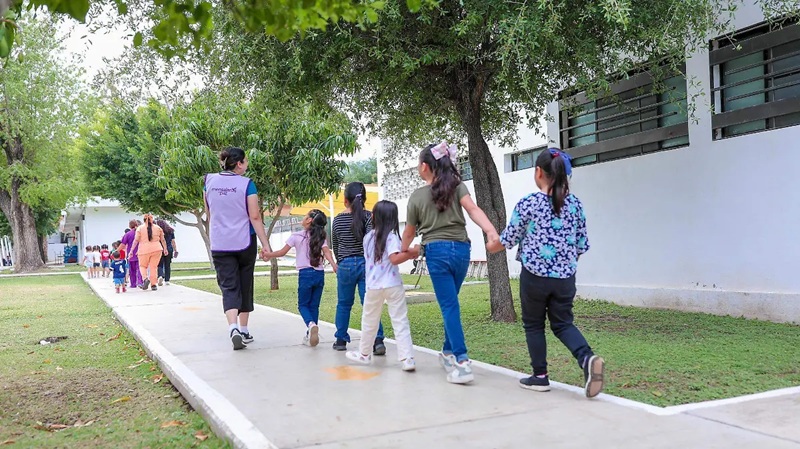 Children holding hands walk in a line along a sunny sidewalk beside trees and a white building.