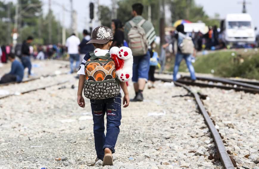 Child wearing a backpack with a teddy bear rides on a railroad track, walking away toward a crowd in the distance.