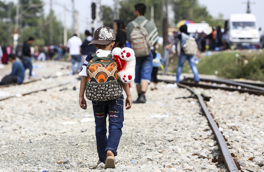 Child wearing a backpack with a teddy bear rides on a railroad track, walking away toward a crowd in the distance.