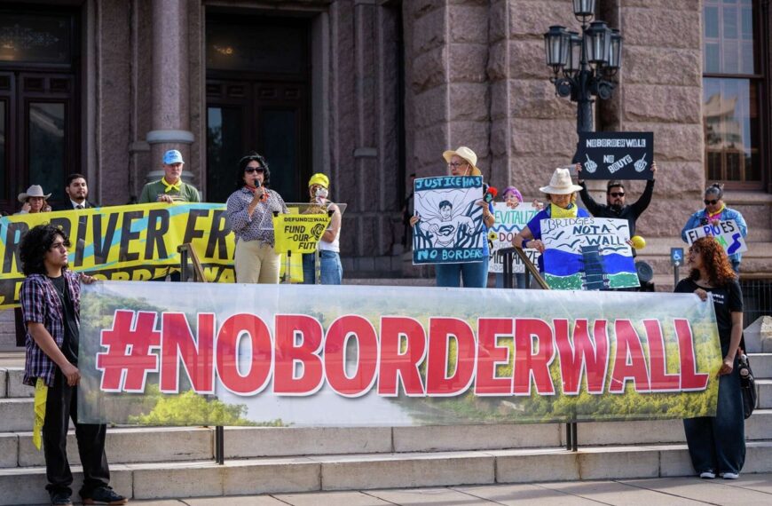 Protesters on a courthouse steps hold a large '#NOBORDERWALL' banner with various protest signs in the background.