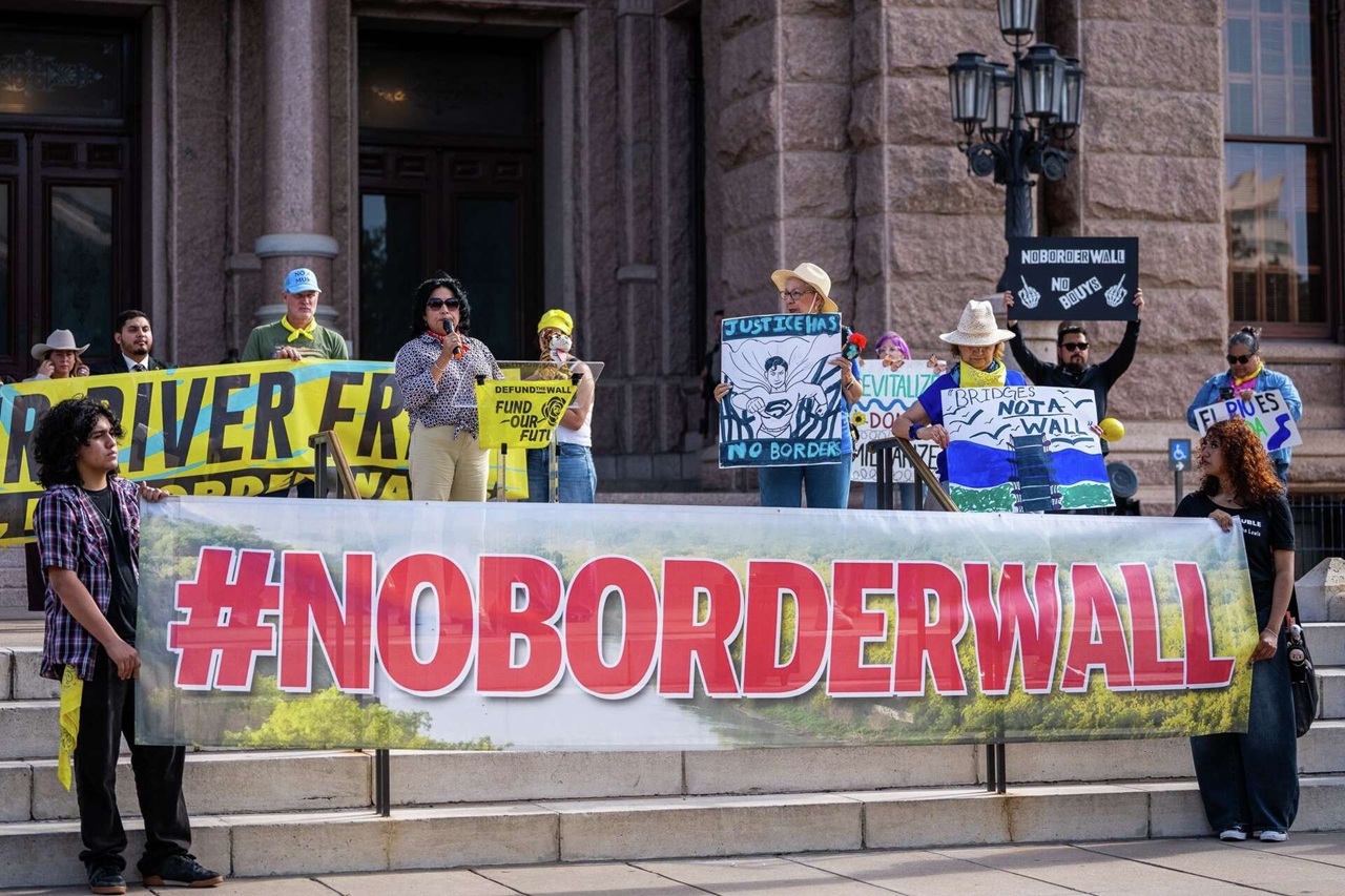 Protesters on a courthouse steps hold a large '#NOBORDERWALL' banner with various protest signs in the background.
