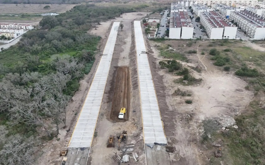 Aerial view of a construction site with two parallel concrete channels and a central dirt trench, heavy machinery at center and apartment buildings in the background.