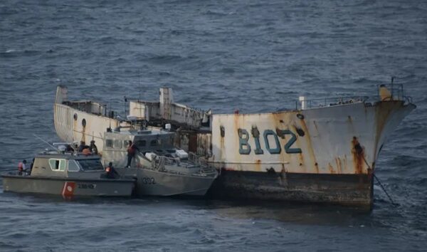 Large, rusted shipwreck with a pale hull partially submerged, surrounded by small rescue boats in open water.