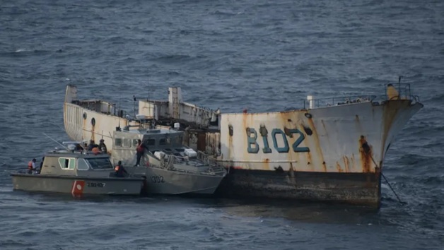 Large, rusted shipwreck with a pale hull partially submerged, surrounded by small rescue boats in open water.