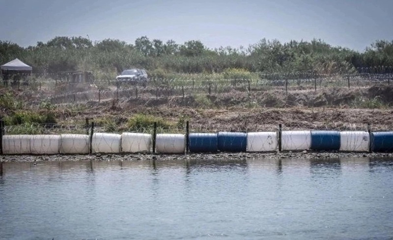 Containment booms float on a calm body of water near a rural shoreline with a car and tent in the background far shore.