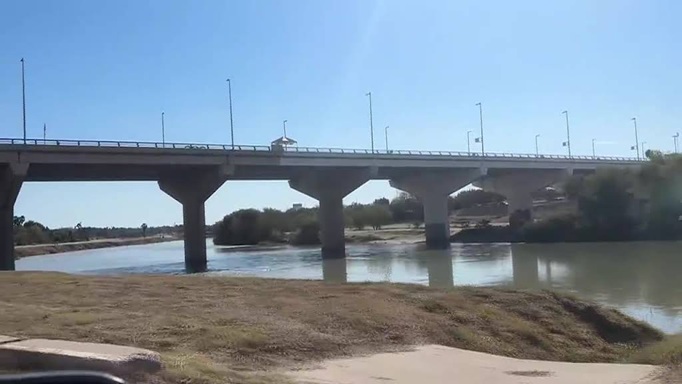 Bridge over a calm river with large concrete piers and streetlights, blue sky above and grassy riverbank in foreground.