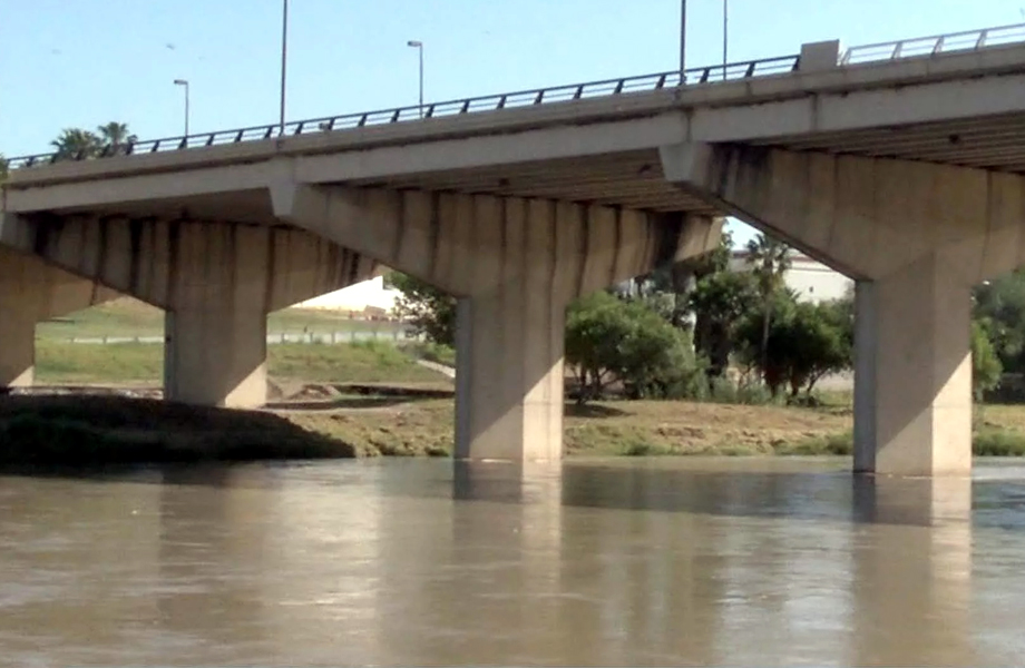 Concrete bridge over a calm river, showing large support pillars and a railing along the top, with trees and grass on the far bank.