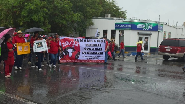 Group of protesters in red clothing marching in the rain, carrying a banner that reads 'Demandamos 40 horas con 2 días de descanso' near a CarneMart store.