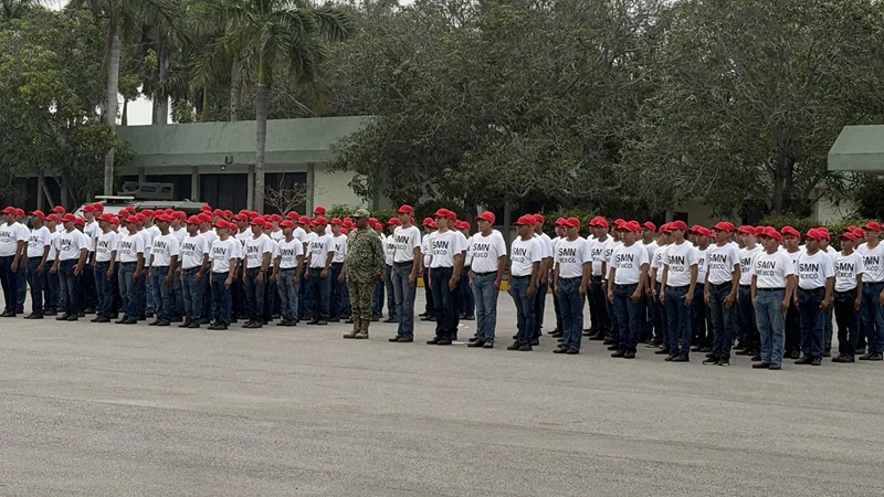 Line of trainees in red caps and white shirts standing at attention outdoors, supervised by a drill instructor.